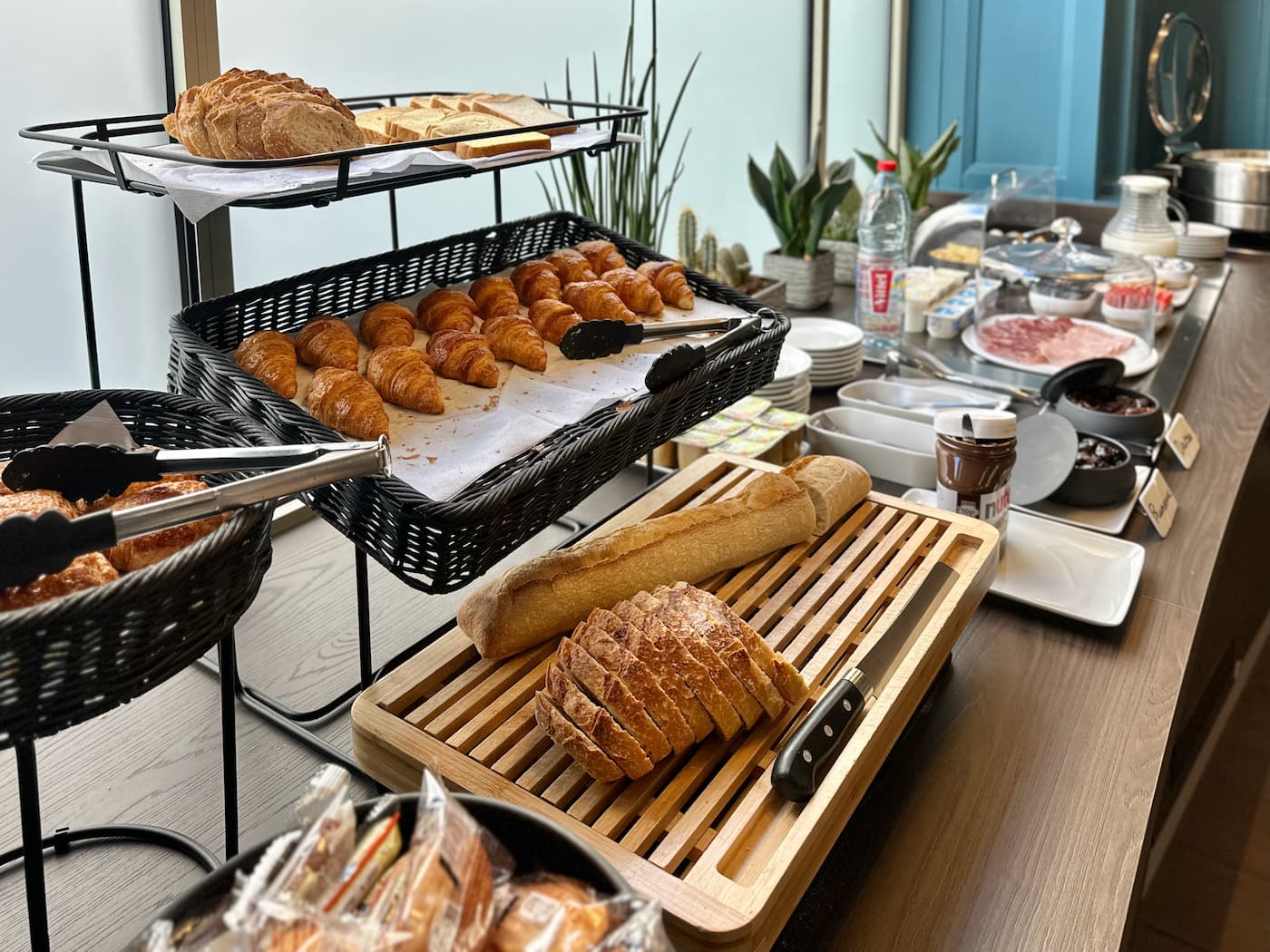 Buffet de petit-déjeuner avec croissants, pain tranché, baguette, Nutella, charcuterie, beurre, et condiments sur une table en bois.