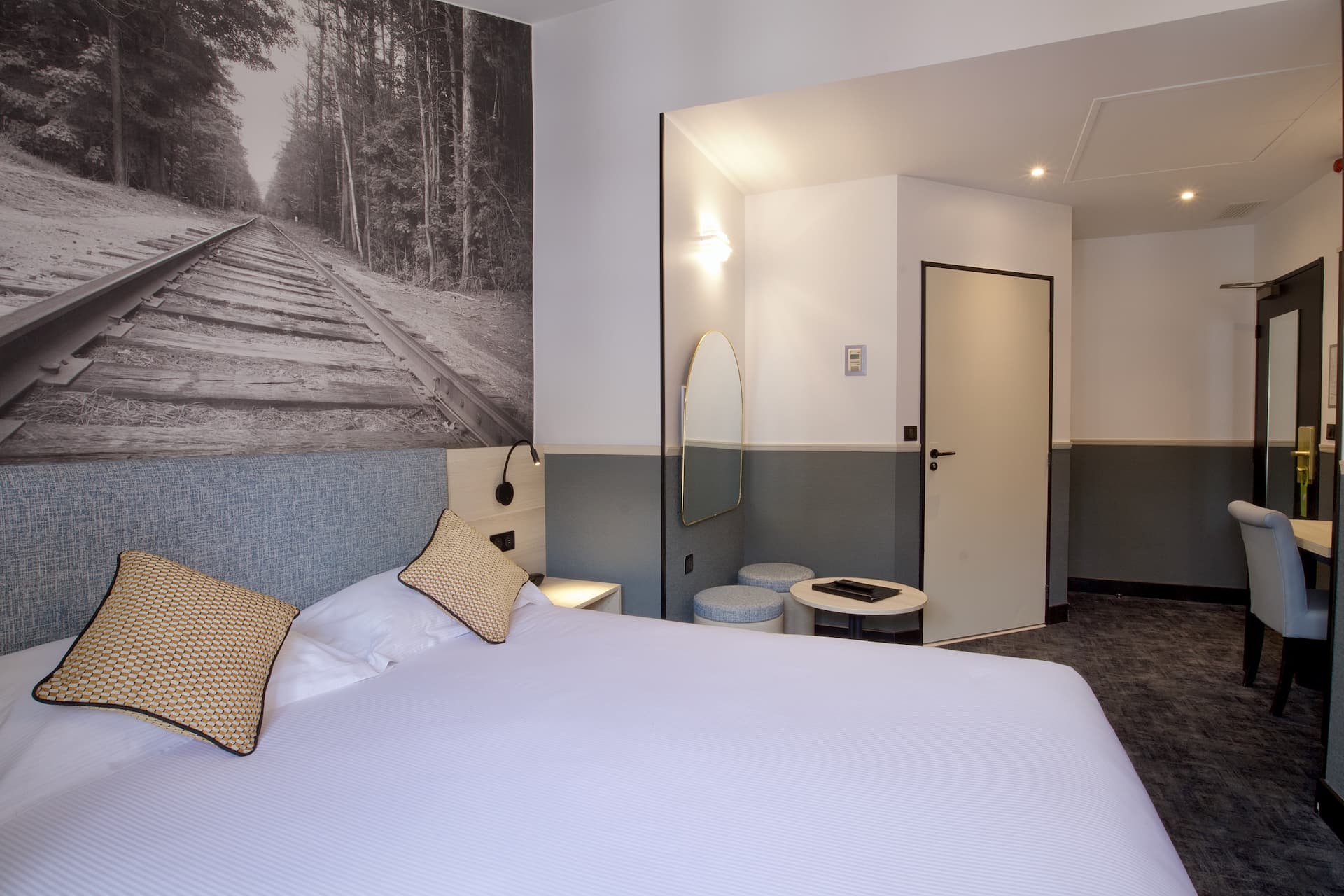 Chambre privilège avec un grand lit blanc, deux coussins beiges, un mur décoré d'une grande photo en noir et blanc de rails de chemin de fer, un miroir ovale, deux tabourets et une petite table.
